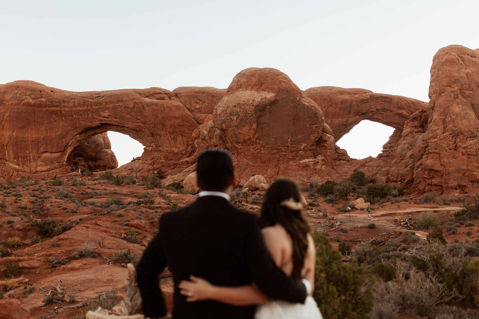 couple stands arm in arm looking at Moab arches