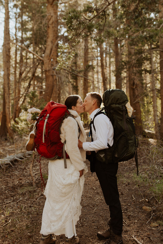 couple kisses wearing backpacks at in woods at outdoor elopement