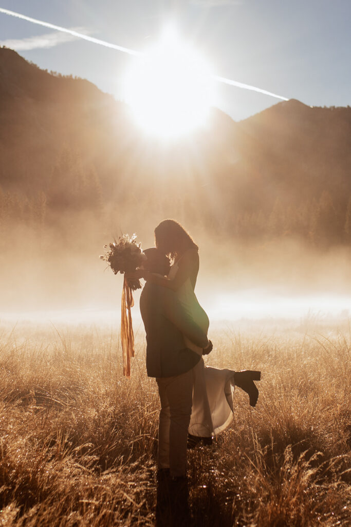 groom lifts bride while she holds wedding bouquet in sunrise