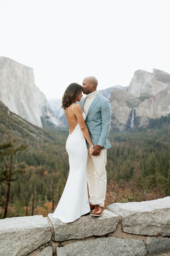 groom kisses bride's forehead standing on ledge at Tunnel View